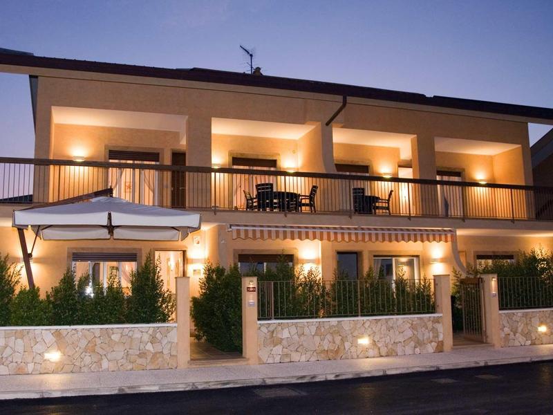 Twee verdiepingen tellend hotel bij schemering met verlichte balkons en een parasol vooraan.