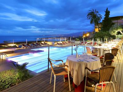 Večerní hotelová terasa se stolkem vedle osvětleného bazénu a výhledem na moře.