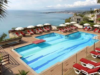 Moderní bazén s červenými lehátky na dřevěné terase s výhledem na moře.