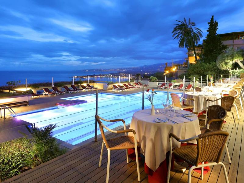 Večerní hotelová terasa se stolkem vedle osvětleného bazénu a výhledem na moře.