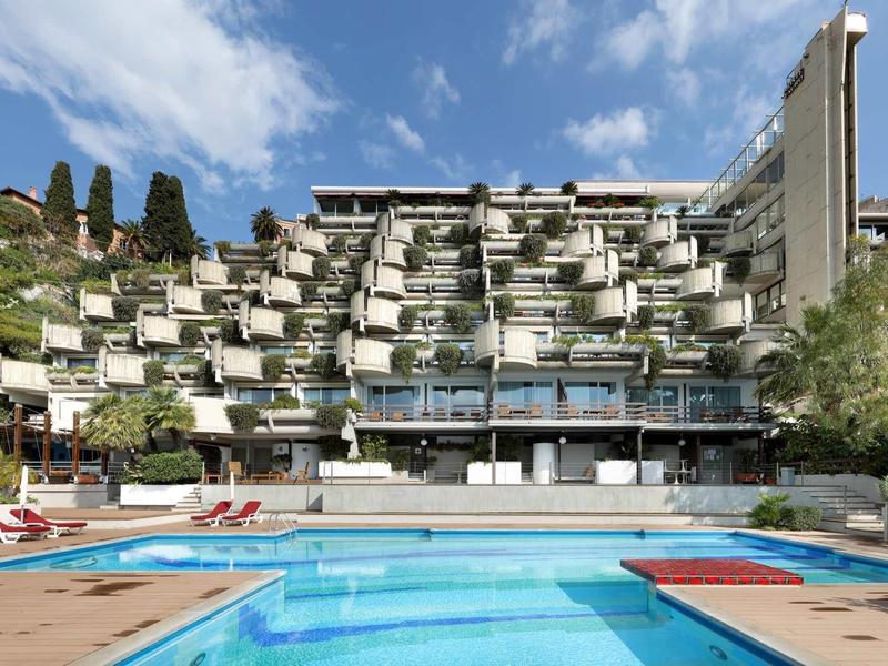 Moderní hotelový komplex na svahu s venkovním bazénem a lehátky pod modrou oblohou.