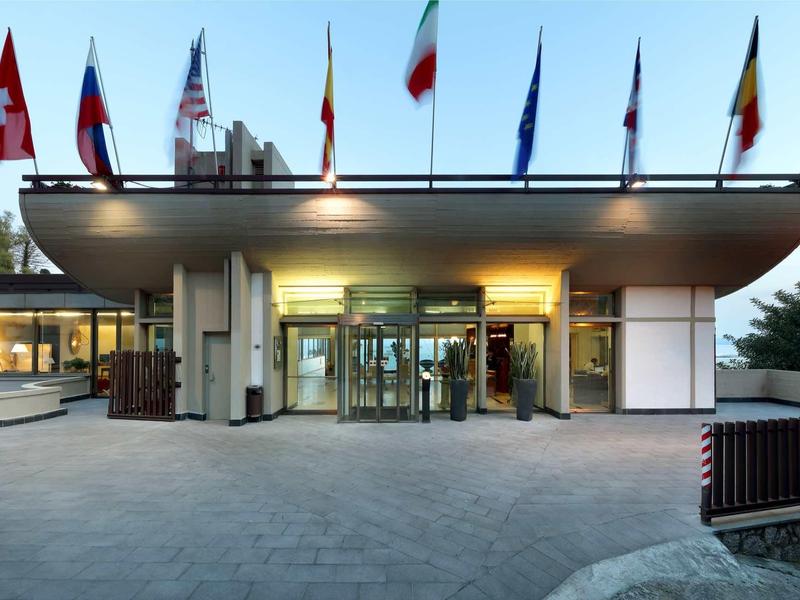 Vchod do moderního hotelu s několika vlajkami na střeše.