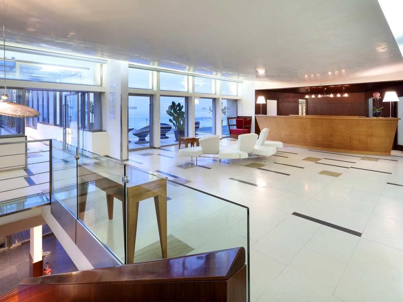 Světlá hotelová hala s recepcí, posezením a velkými okny.