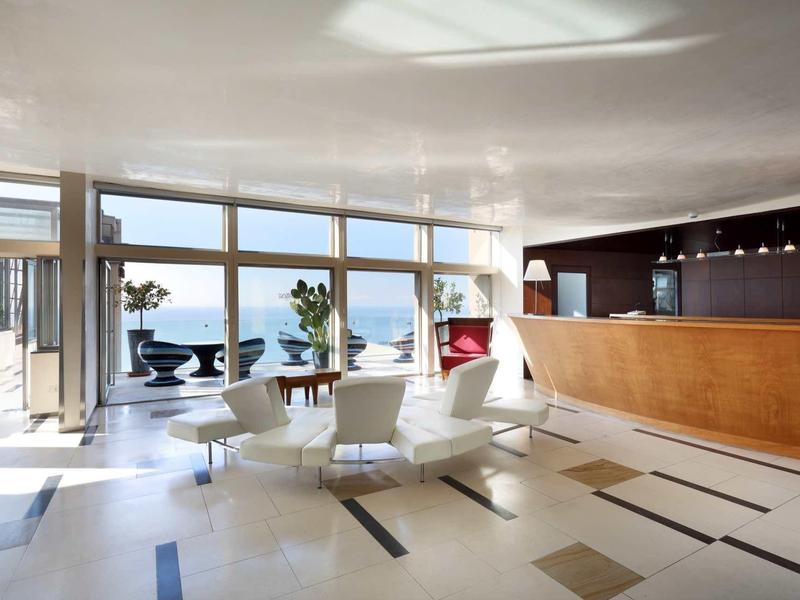 Světle hotelové lobby s velkými okny, bílým posezením a výhledem na moře.