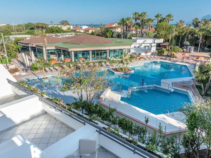 Hotelový bazén s lehátky a slunečníky obklopený palmami a budovami pod jasnou oblohou.