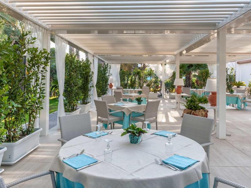 Terasa restaurace s kulatými stoly, bílými ubrusy a modrými polštářky na židlích pod pergolou.