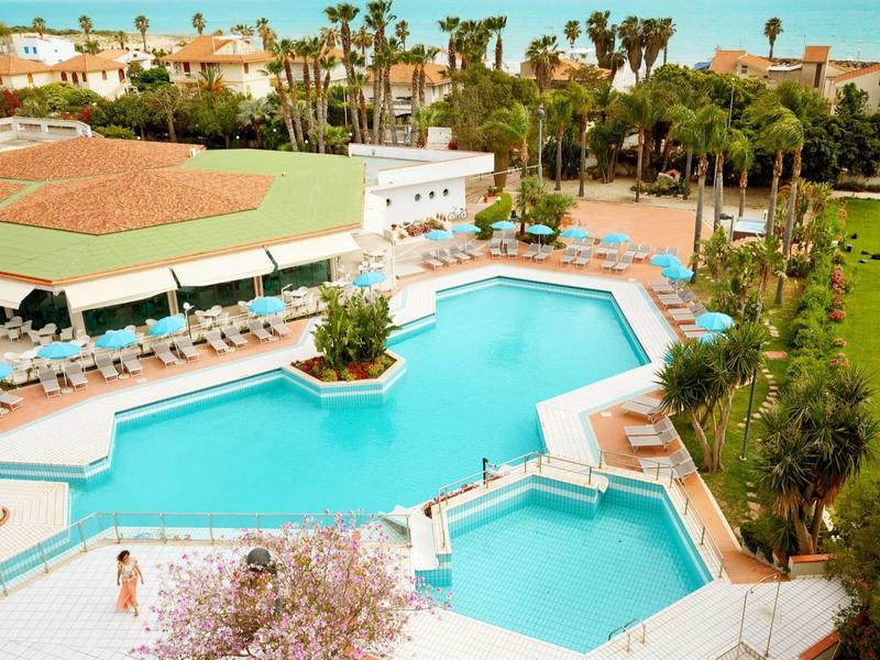 Hotelový bazén s lehátky, obklopený palmami a budovami za slunečného nebe.