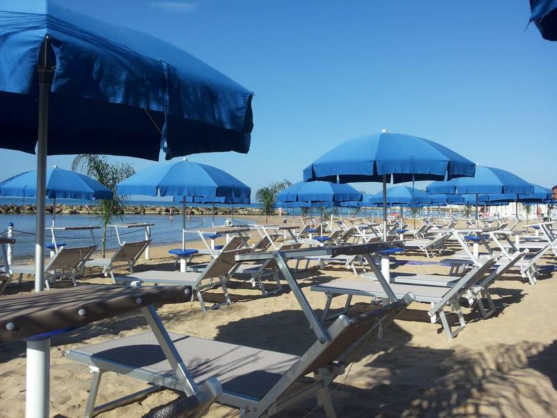 Prázdná lehátka a modré slunečníky na písčité pláži pod jasnou oblohou.