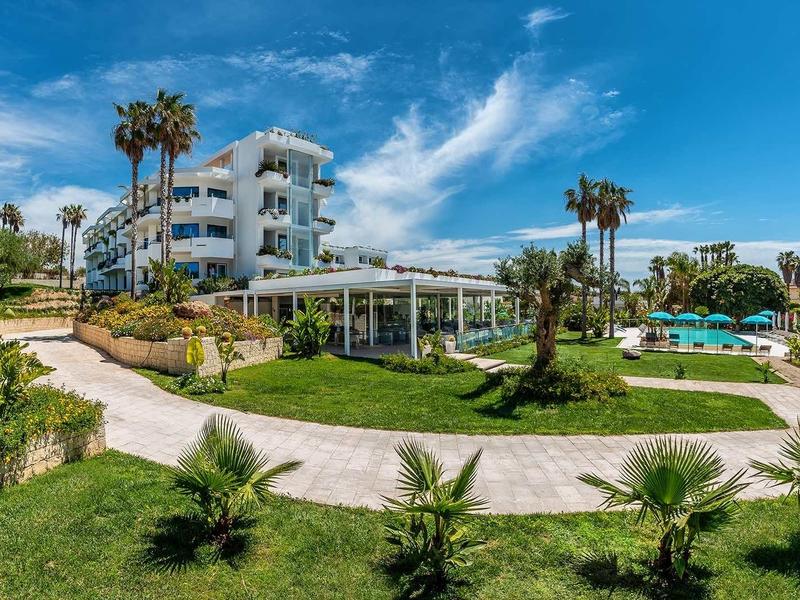 Moderní hotelový komplex s bazénem, palmami a upravenou trávou pod modrou oblohou.