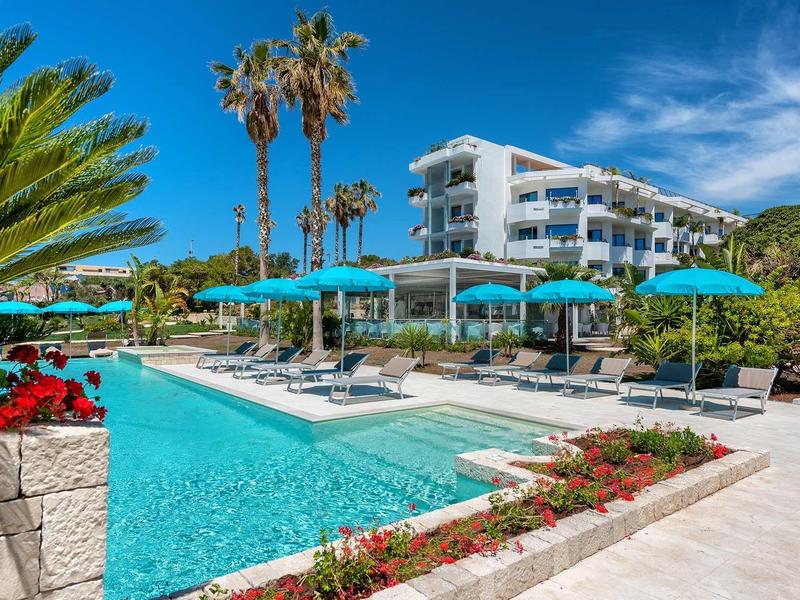 Moderní venkovní bazén s lehátky, slunečníky a palmami pod jasnou oblohou.