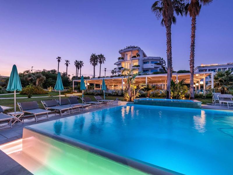 Osvětlený hotel s bazénem, lehátky a palmami za soumraku.