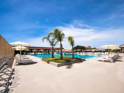 Ampia area piscina esterna con lettini, ombrelloni e palme sotto un cielo azzurro.