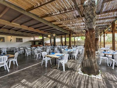 Duża zadaszona przestrzeń na zewnątrz z białymi plastikowymi krzesłami i stołami, drzewo na środku.