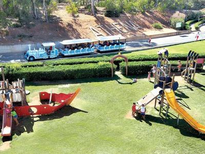 Plac zabaw dla dzieci z zamkiem z piasku, zjeżdżalnią i pluszakiem na zielonej trawie obok mini pociągu.