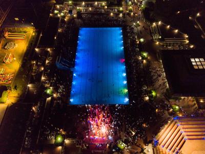 Vista notturna di una piscina rettangolare illuminata circondata da persone e luci.