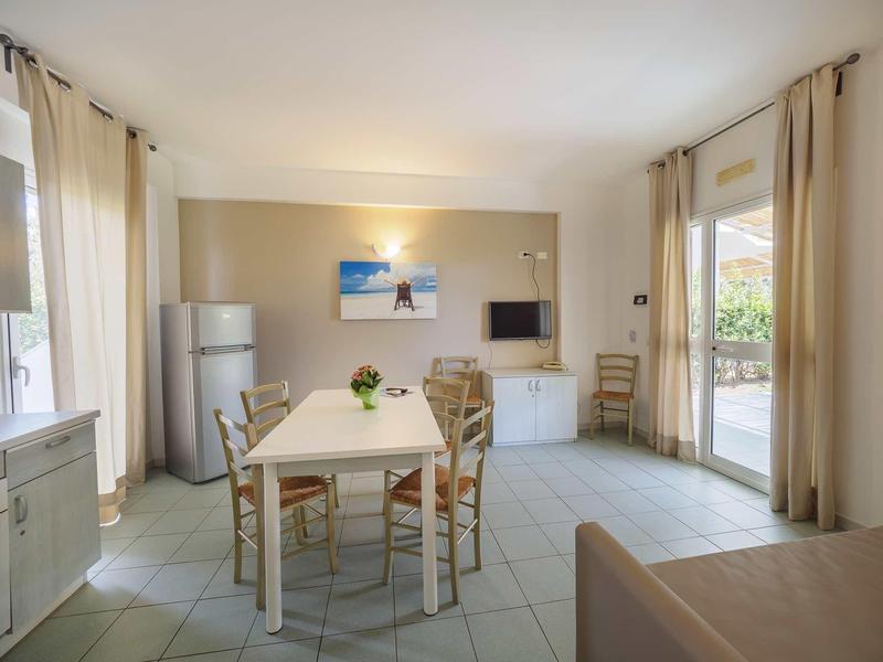 Jasna jadalnia z białym stołem, krzesłami, lodówką, telewizorem i dużymi oknami z zasłonami.