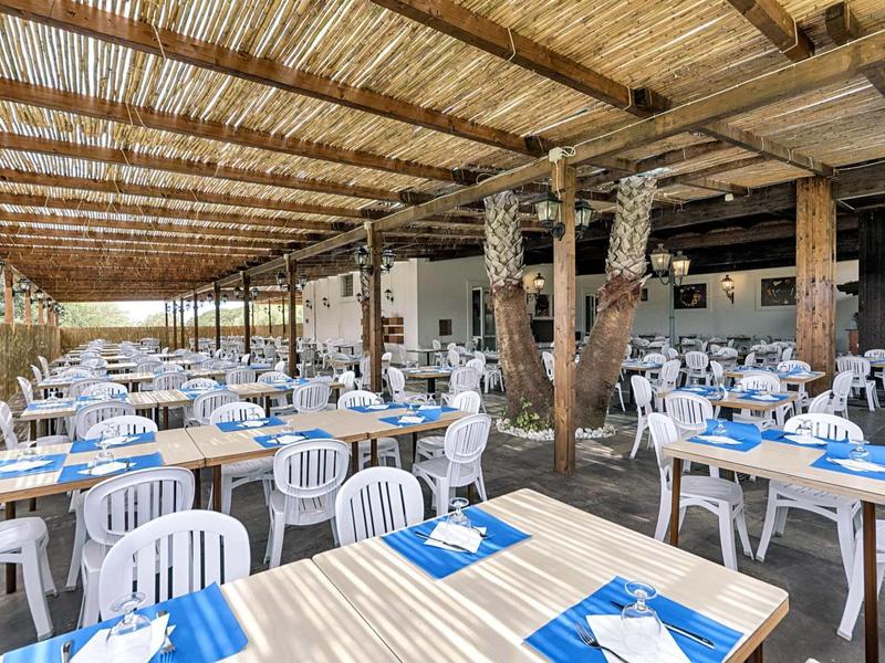 Duży zewnętrzny obszar restauracji z białymi plastikowymi krzesłami i niebieskimi podkładkami pod talerze pod dachówką ze słomy.