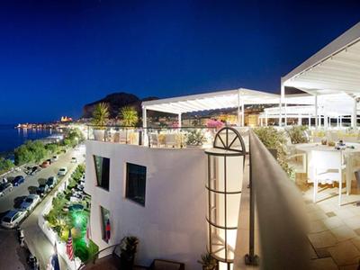 Vue en soirée d'une terrasse de restaurant ouverte avec tables et chaises, surplombant une route côtière et la mer.