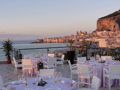 Terrasse avec tables dressées, vue sur ville côtière et mer au coucher du soleil.