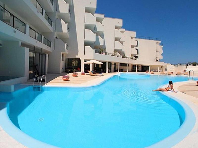Grande piscine bleu clair devant un hôtel blanc sous un ciel bleu clair.