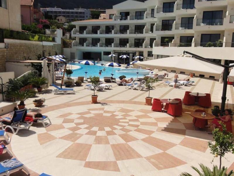 Complexe hôtelier avec piscine, transats et zones ombragées sous un ciel bleu.