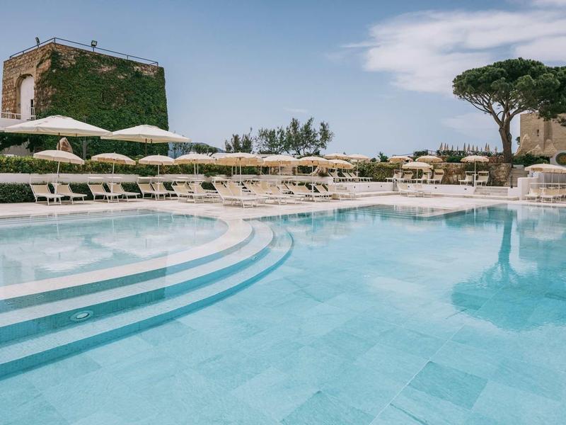 Large hotel pool with sun loungers and umbrellas under a blue sky.