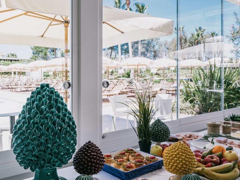 Hell gedeckter Tisch mit buntem Gebäck und Obst vor großer Fensterfront mit Blick ins Freie.