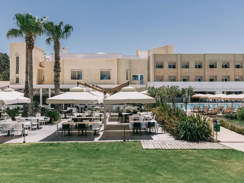 Moderner Gebäudekomplex mit Sonnenschirmen, Sitzgruppen und Palmen vor grünem Rasen unter blauem Himmel.