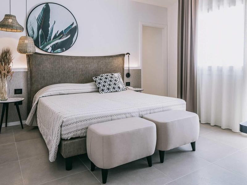 Modernes Schlafzimmer mit grauem Bett, zwei grauen Hockern, rundem Kunstwerk und großem Fenster.