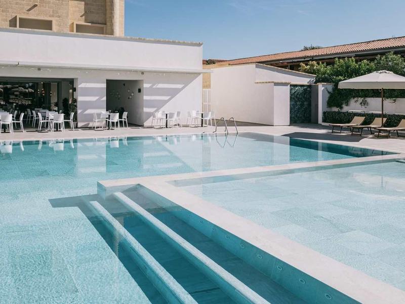 Moderner Außenpool mit klar blauem Wasser, weißen Stufen und angrenzenden Gebäuden bei klarem Himmel.