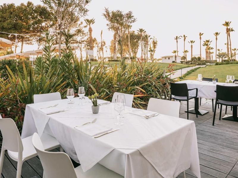 Weiß gedeckter Esstisch auf Terrasse mit Stühlen, grünen Pflanzen und Palmen im Hintergrund.