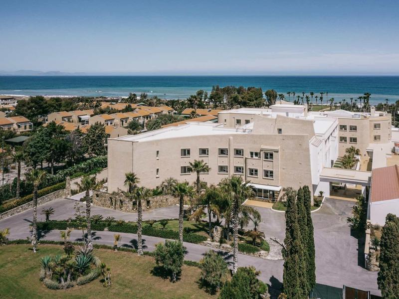 Große weiße Gebäudekomplex mit grünen Palmen und blauem Meer im Hintergrund unter klarem Himmel.