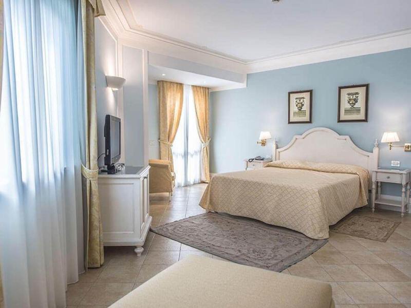 Chambre d'hôtel lumineuse avec lit double, tables de nuit, télévision et grande fenêtre avec rideaux.