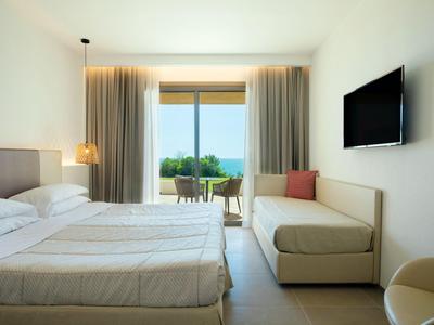 Světý hotelový pokoj s manželskou postelí, pohovkou, televizí a balkonem s výhledem na moře.