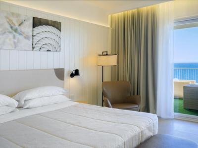 Světle zařízený hotelový pokoj s manželskou postelí, křeslem, stojací lampou a výhledem na moře skrz skleněná okna.