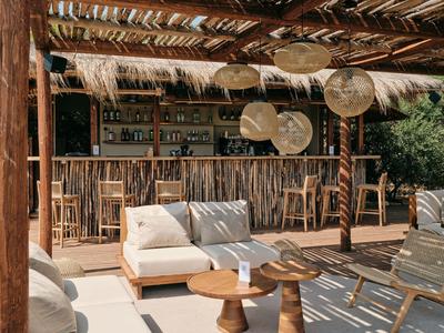 Otevřený dřevěný bar s barovými židličkami a útulným posezením pod dřevěnou a slámovou střechou.