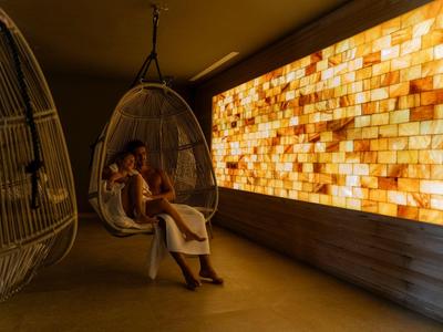 Dva lidé relaxují v závěsných křeslech před osvětlenou stěnou z krystalů soli v relaxačním prostoru.