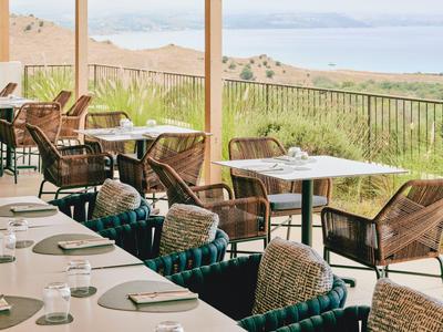 Venkovní restaurace s ratanovými židlemi a stoly s výhledem na moře a kopce.