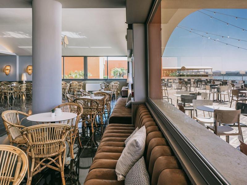 Vnitřní část restaurace s ratanovými židlemi a venkovní část s výhledem na moře za slunečného světla.