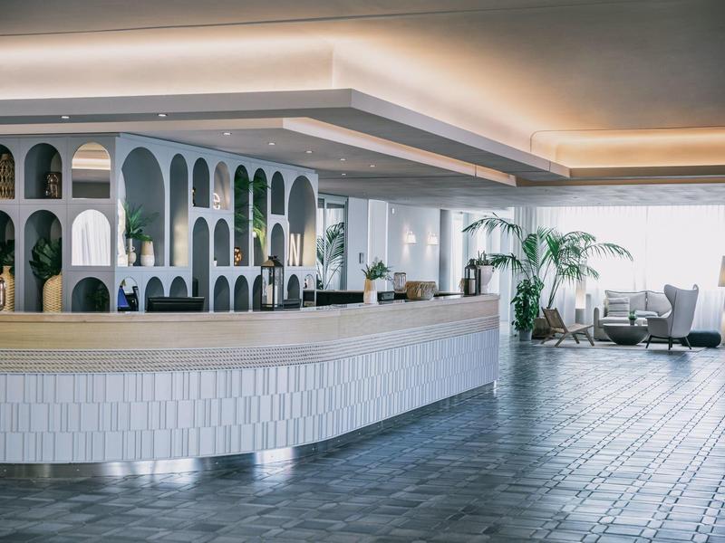 Moderní hotelové lobby s zakřivenou recepcí a velkými okny vpouštějícími denní světlo.