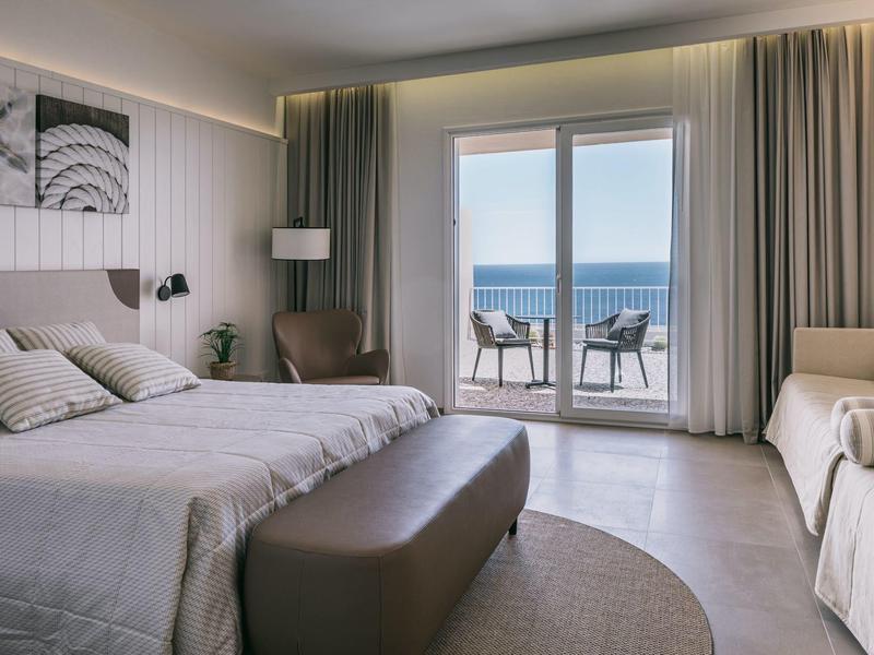 Moderní hotelový pokoj s manželskou postelí, posezením a terasou s výhledem na moře.