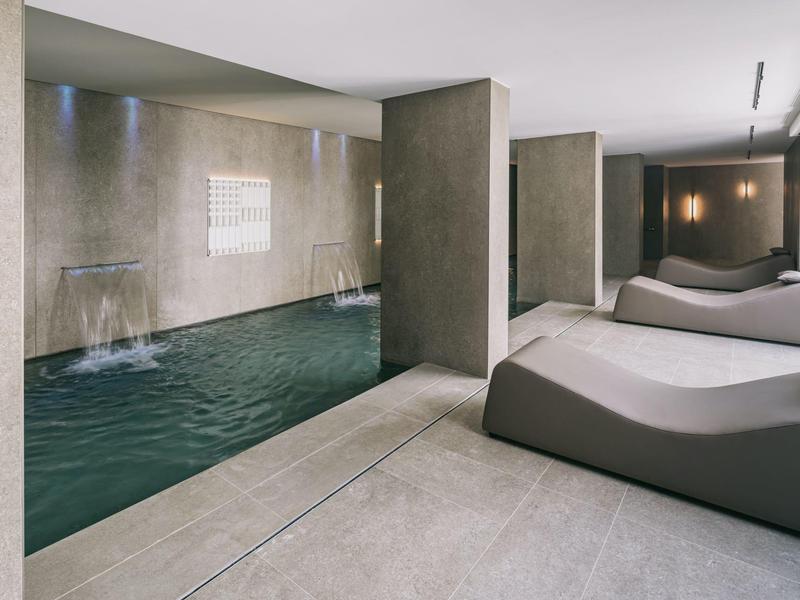 Moderní wellness prostor s bazénem, vodopády a lehátky v neutrálních odstínech.