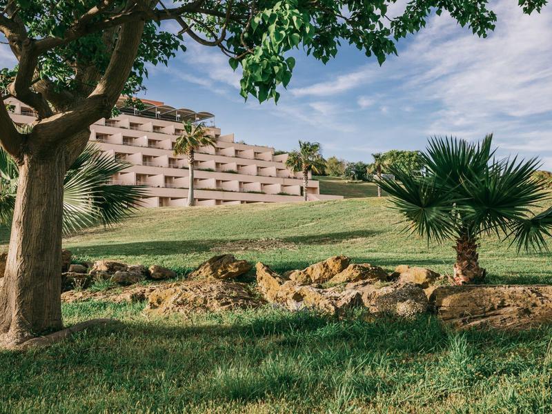 Moderní hotelová budova s balkony, obklopená zelenou trávou a palmami pod modrou oblohou.