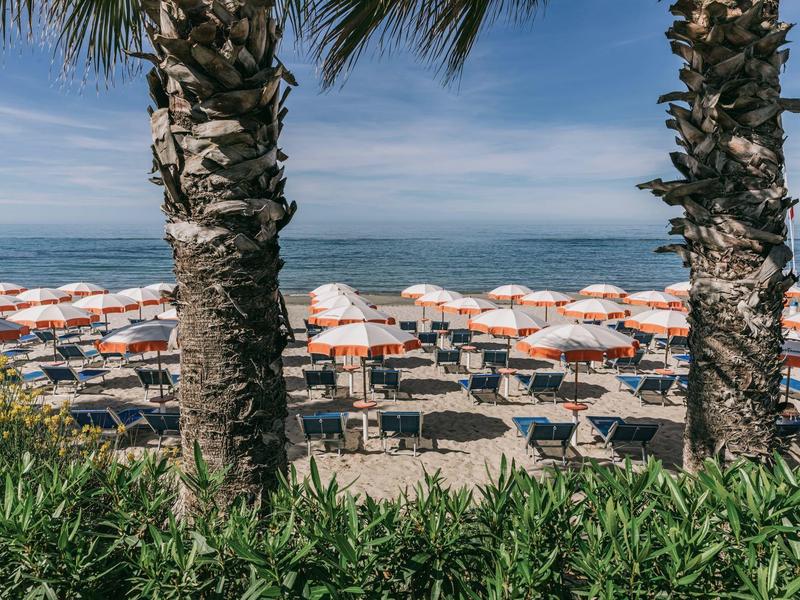 Pláž s řadami slunečníků a lehátek mezi palmami a zelenými keři.