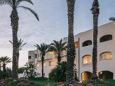 Mehrstöckiges weißes Gebäude mit gebogenen Fenstern, umgeben von hohen Palmen und grünem Rasen.