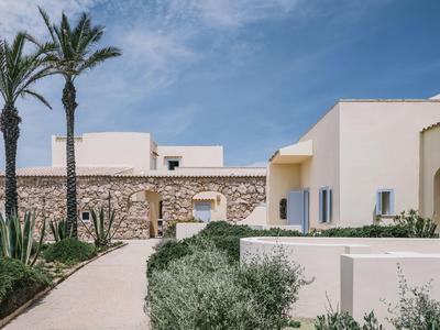 Moderní hotelový komplex s bílými stěnami, palmami a kamennými prvky pod modrou oblohou.