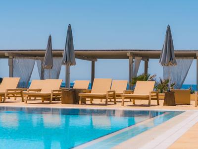 Bazén s lehátky a zavřenými slunečníky na pozadí čisté oblohy a výhledu na moře.