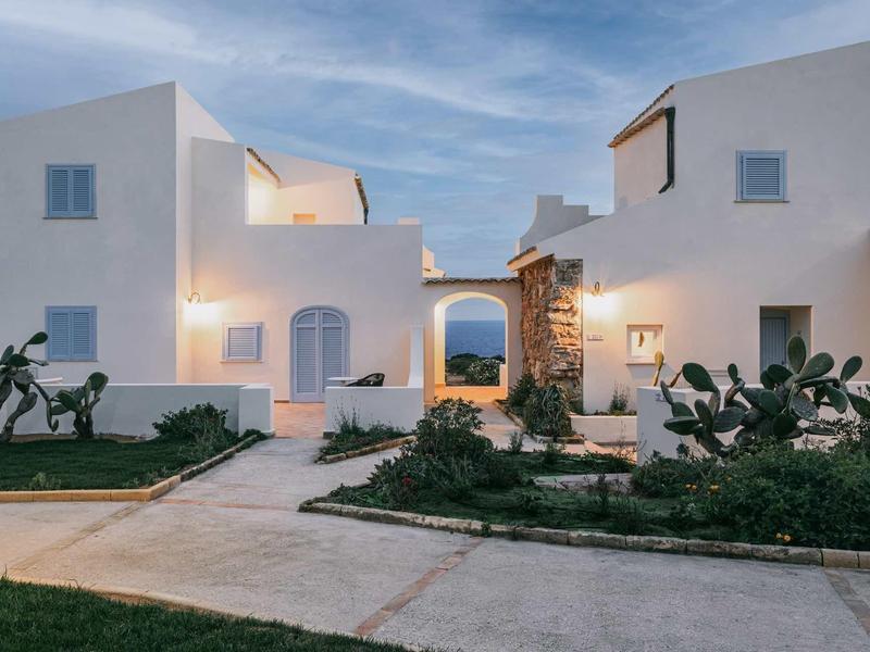 Witgekalkte gebouwen met vierkante ramen en verzorgde tuinpaden onder een blauwe lucht.