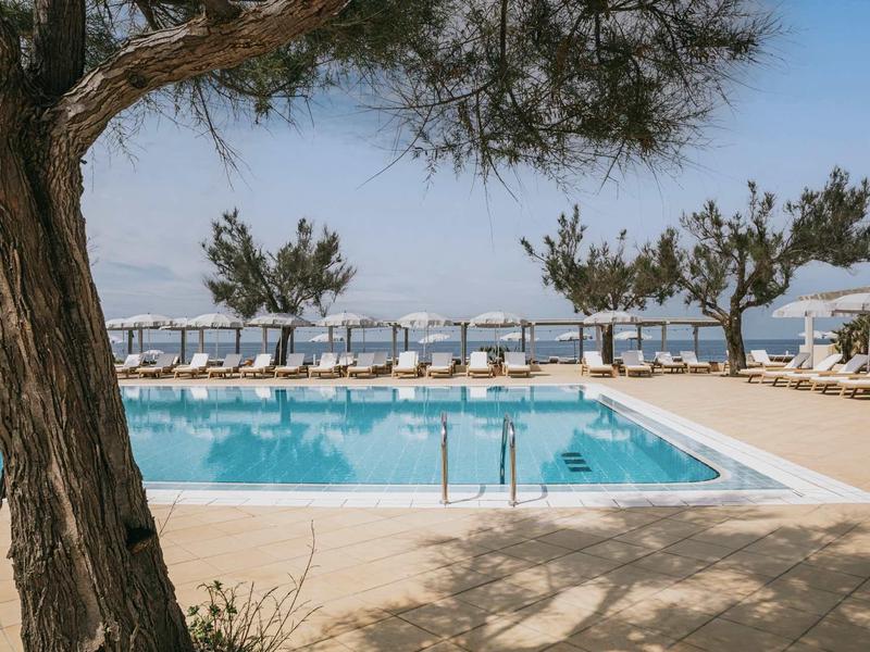 Helder zwembad omgeven door ligstoelen en bomen met uitzicht op de zee.