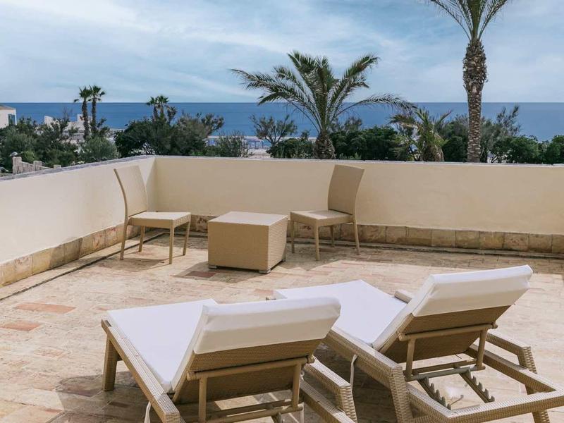 Terras met twee ligstoelen met uitzicht op zee en palmbomen onder een bewolkte lucht.